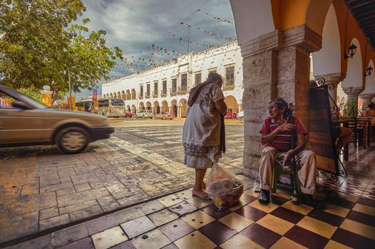 Valladolid, Mexico 20 august 2022: Old beggar of the Mayan ethnic group is sitting under the porticoes asking for alms while all around him runs in a frenetic way: Example of classism in the Mexican city of Valladolid. Street side beggars