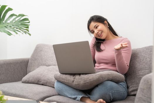 Happy young asian woman calling with mobile phone and usin laptop computer on sofa at home.