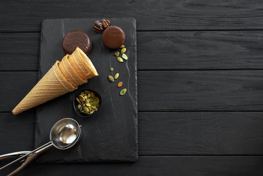 Ingredients for Cookies N' Cream ice cream with chocolate and cookies, nuts, sugar cones, tubs and a metal scoop scattered on a slate, top view. Copy space