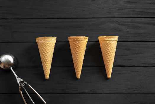 Three waffle cups for ice cream empty on a black wooden background view from above. Next to the ice cream scoop. Top view