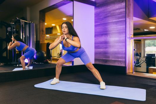 young beautiful sports woman in the gym doing exercises on a mat, in front of a mirror, arm swing, squats, fitness, yoga, in sportswear