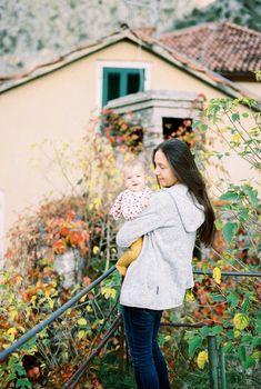 Mom with a baby in her arms stands on the balcony of the house near the tree. High quality photo