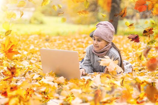 Cute little girl holds an orange maple leaf near the face. Pretty blond girl wearing beige coat, knitted cap and scarf has fun in autumn park, close-up. High quality photo