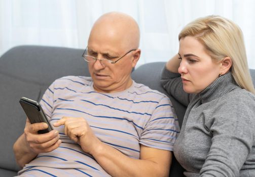 Smiling older father with adult daughter using phone together, having fun, watching video in social network or shopping online, caring young woman teaching mature dad or grandfather to use smartphone