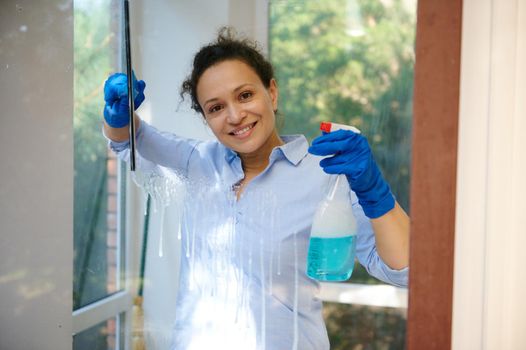 Multi-ethnic woman with blue rubber gloves, smiles looking at the camera while washing windows with a detergent and glass cleaning scraper, removing streaks and stains. Household chore, housekeeping
