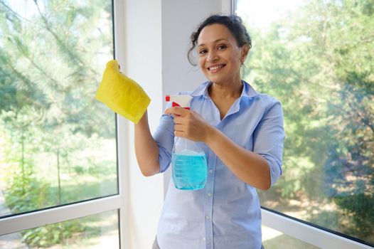 Hispanic pretty woman in blue shirt, maid, housewife sprays glass cleaner detergent on yellow cloth, smiles looking at camera, standing against the veranda panoramic windows during the spring cleaning