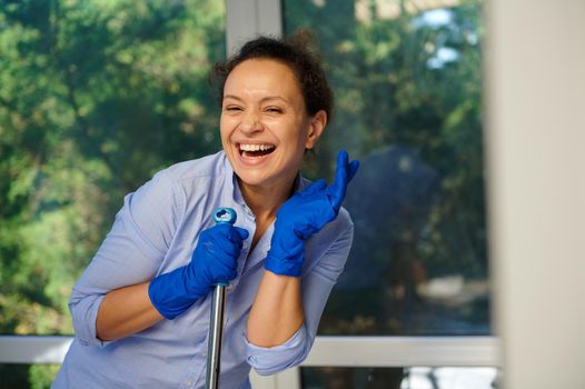 Housekeeping Job Concept. Stunning Latin American woman in casual blue shirt and rubber work gloves happily doing homework cleaning routine, posing with a mop and cutely smiling looking at camera