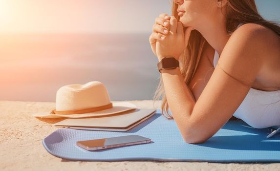 Successful business woman in yellow hat working on laptop by the sea. Pretty lady typing on computer at summer day outdoors. Freelance, travel and holidays concept.