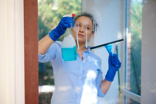 View through a glass of a pretty housewife spraying glass cleaner detergent on the windows and using a scraper to remove stains, streaks and dust during spring cleaning. Household chores. Housekeeping