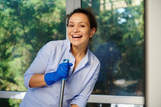 Close-up portrait. Beautiful woman in blue shirt and rubber gloves, holding mop, smiling cheerful toothy smile, looking at camera, happily doing household chores, mopping floor, disinfecting surfaces