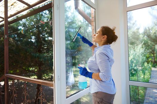 Waist-up portrait of pretty woman in rubber gloves cleaning window with detergent at home. Lovely housewife keeping her home tidy, happily performing domestic chores, enjoying housework. Housekeeping