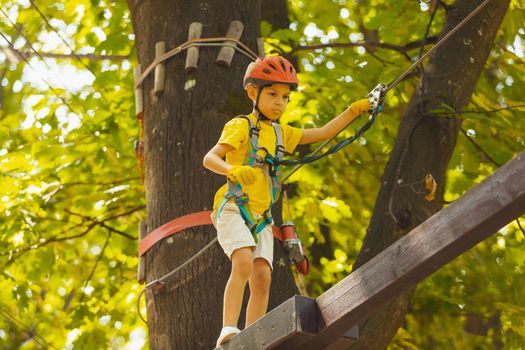 Portrait of a concentrated boy who is climbing in the rope park in summer. He is wearing a protective helmet and gloves