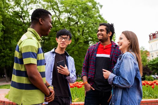 Joyful smiling students during outdoor weekend meeting. Lovely girl in denim shirt and three multiethnic boys in casual clothes talking together outdoors at the campus area.