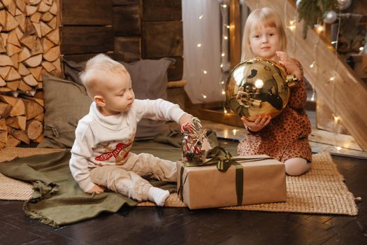 Little brother and sister play on Christmas eve in a beautiful house decorated for the New Year holidays. Children are playing with a Christmas gift. Scandinavian-style interior with live fir trees and a wooden staircase.