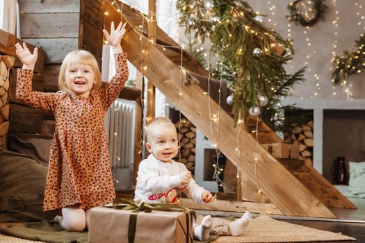 Little brother and sister play on Christmas eve in a beautiful house decorated for the New Year holidays. Children are playing with a Christmas gift. Scandinavian-style interior with live fir trees and a wooden staircase.