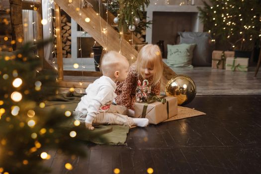 Little brother and sister play on Christmas eve in a beautiful house decorated for the New Year holidays. Children are playing with a Christmas gift. Scandinavian-style interior with live fir trees and a wooden staircase.