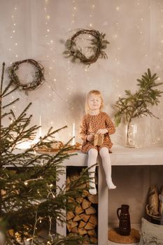 A girl is playing on Christmas Eve in a beautiful house decorated for the New Year holidays. Scandinavian-style interior with live fir trees and a wooden staircase.