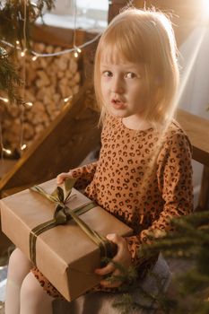 A little blonde girl is sitting on a wooden staircase in a Scandinavian interior decorated in a New Year's style. A child holds a Christmas gift in a craft package.