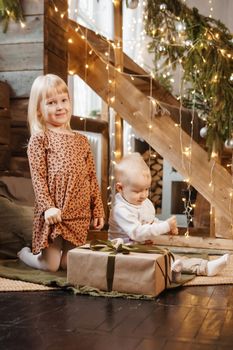 Little brother and sister play on Christmas eve in a beautiful house decorated for the New Year holidays. Children are playing with a Christmas gift. Scandinavian-style interior with live fir trees and a wooden staircase.