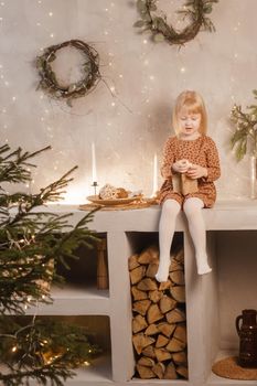 A girl is playing on Christmas Eve in a beautiful house decorated for the New Year holidays. Scandinavian-style interior with live fir trees and a wooden staircase.