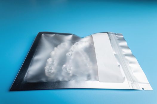 Silver plastic package body with transparent braces inside on a blue background. Invisible removable retainers for orthodontic treatment.