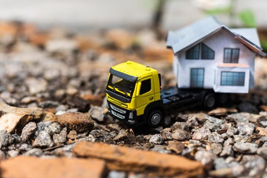 Close-up of a small toy truck on a tree stump carrying a miniature house model on top against a blurred forest background, front view. Countryside relocation service concept