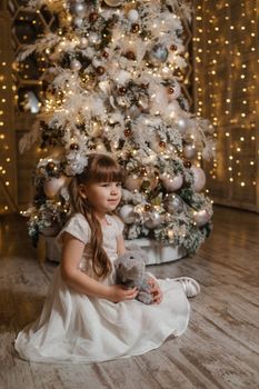 A girl in a festive light dress with a plush toy in her hands next to a Christmas tree, lights of garlands in the background. The concept of New Year holidays.