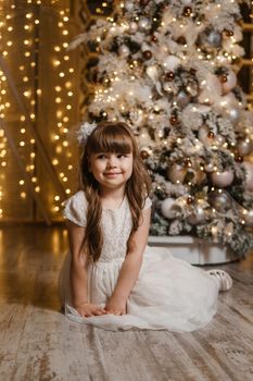 A girl in a festive light dress next to a Christmas tree, lights of garlands in the background. The concept of New Year holidays.