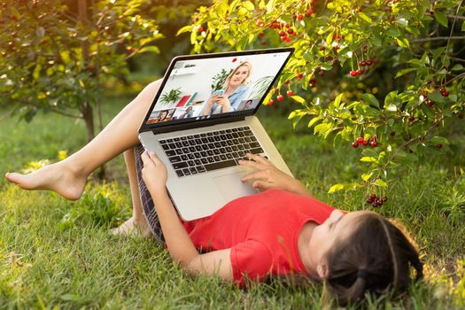 Beautiful young lady is chatting with friends outdoors as it is very useful to be in nature. Laptop is the best friend for a child nowadays. Green and vast meadow.