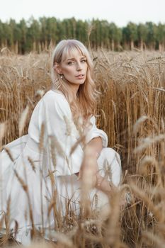 A blonde woman in a long white dress walks in a wheat field. The concept of a wedding and walking in nature.