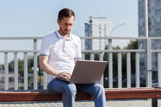 An adult man works on his laptop while sitting on a bench. The concept of remote work. The writer works remotely, enjoying nature.