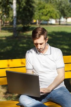 An adult man works on his laptop while sitting on a bench. The concept of remote work. The writer works remotely, enjoying nature.