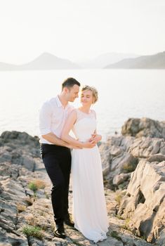 Groom hugs bride from behind standing on the rocks near the sea. High quality photo