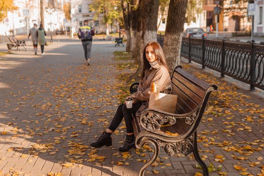A stylish brunette woman walks around the autumn city. The brunette is sitting on a bench outside with a glass of coffee and a phone, holding a craft bag with a baguette.