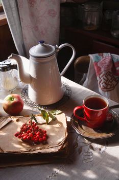 Red cup of tea on vintage metal plate with a bunch of red rowan lying on old book and red apple in the table in rustic kitchen, autumn morning tea concept, selective focus.