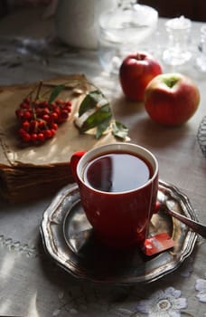Red cup of tea on vintage metal plate with a bunch of red rowan lying on an old book and a couple of red apples, autumn morning tea concept, selective focus