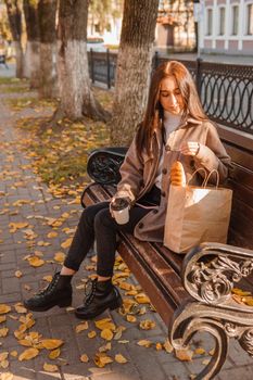 A stylish brunette woman walks around the autumn city. The brunette is sitting on a bench outside with a glass of coffee and a phone, holding a craft bag with a baguette.