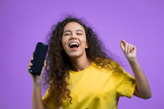 Mixed race woman listening to music by wireless portable speaker - modern sound system. Teenager dancing, enjoying on violet studio background. She moves to the rhythm of music. High quality photo
