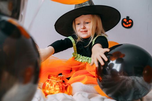 Children's Halloween - a girl in a witch hat and a carnival costume with airy orange and black balloons at home. Ready to celebrate Halloween.