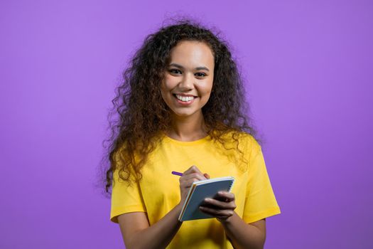 Mixed race woman making notes in planner, lady holding pen. She writes future plans and to-do list in notebook for week or month. Keeping personal diary on purple studio background. High quality photo