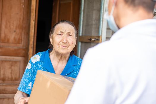 Young male volunteer in mask gives an elderly woman boxes with food near her house. The son helps a single elderly mother. Family support, caring. Quarantined, isolated. Coronavirus covid-19. Donation
