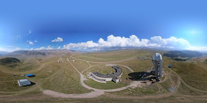 360 degree photo of the Assy-Turgen Observatory. A tall building with a dome and a telescope. Several houses and a tent camp. Big white clouds, mountains and hills covered with yellow-green grass