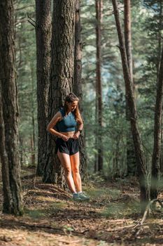 Young woman in sportswear leaned on a tree and straightens clothes in the forest during working out. Trail running and active life concept