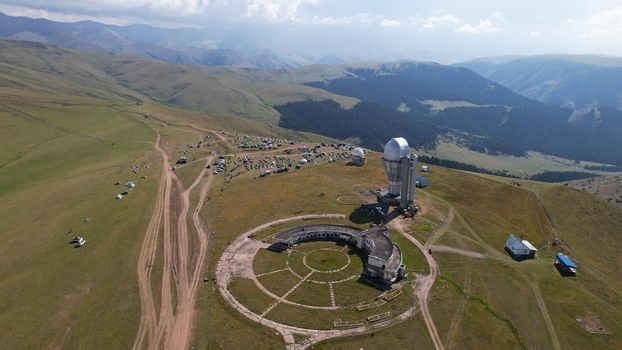 The Assy-Turgen Observatory is high in the mountains. There is a tent camp next to the observatory. Large cumulus clouds in a blue sky. Yellow-green hills, forest in places. Top view from a drone