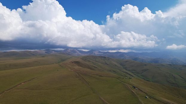 Big white clouds over green hills and mountains. Top view from the drone on endless fields. Roads are visible in places, herds of animals graze. A tent camp has been set up. Coniferous trees in gorge