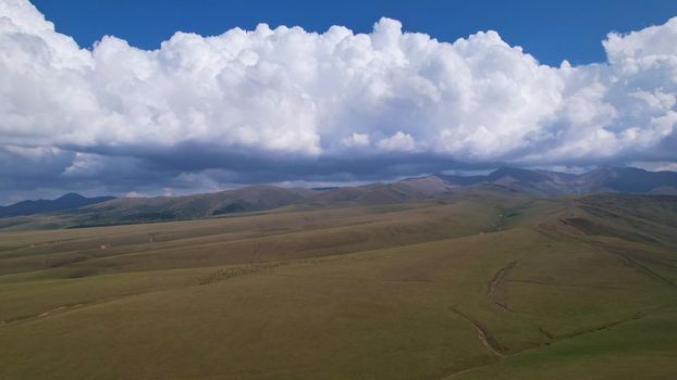 Big white clouds over green hills and mountains. Top view from the drone on endless fields. Roads are visible in places, herds of animals graze. A tent camp has been set up. Coniferous trees in gorge