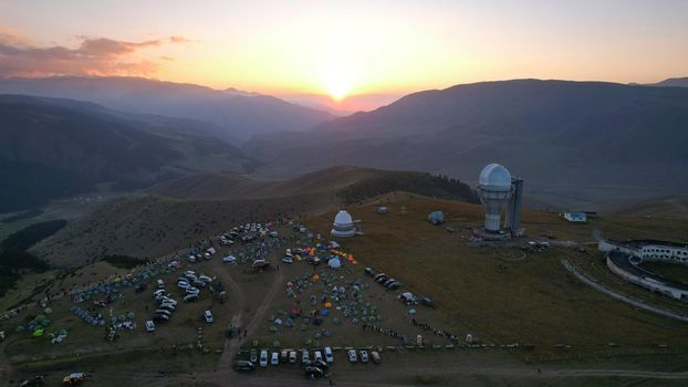 Two large telescope domes at sunset. Drone view of Assy-Turgen Observatory. Beautiful red sunset. Green hills and clouds. Tourists watch the sun. There is a large tent camp and cars nearby. Kazakhstan