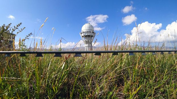 A large observatory is protected by a barbed fence. There is a long sharp wire around perimeter. Buildings with domes and a telescope are visible in distance. Yellow-green grass. Mountainous terrain