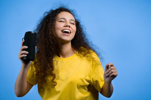 Mixed race woman listening to music by wireless portable speaker - modern sound system. Teenager dancing, enjoying on blue studio background. She moves to the rhythm of music. High quality photo