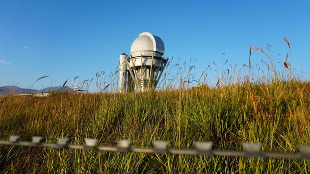 A large observatory is protected by a barbed fence. There is a long sharp wire around perimeter. Buildings with domes and a telescope are visible in distance. Yellow-green grass. Mountainous terrain